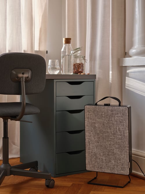 A black FÖRNUFTIG air purifier standing on the floor beneath a study space with a dark-grey BLECKBERGET swivel chair.