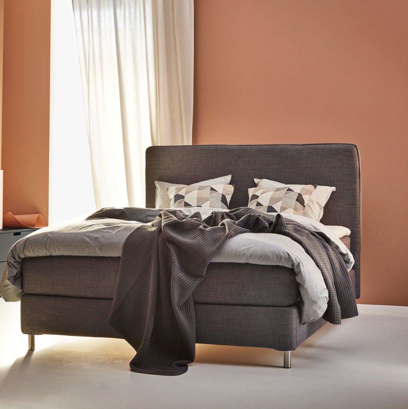 Bäddad säng med grå textilier, geometriska kuddar, persikofärgad vägg och fönster med vita gardiner.
