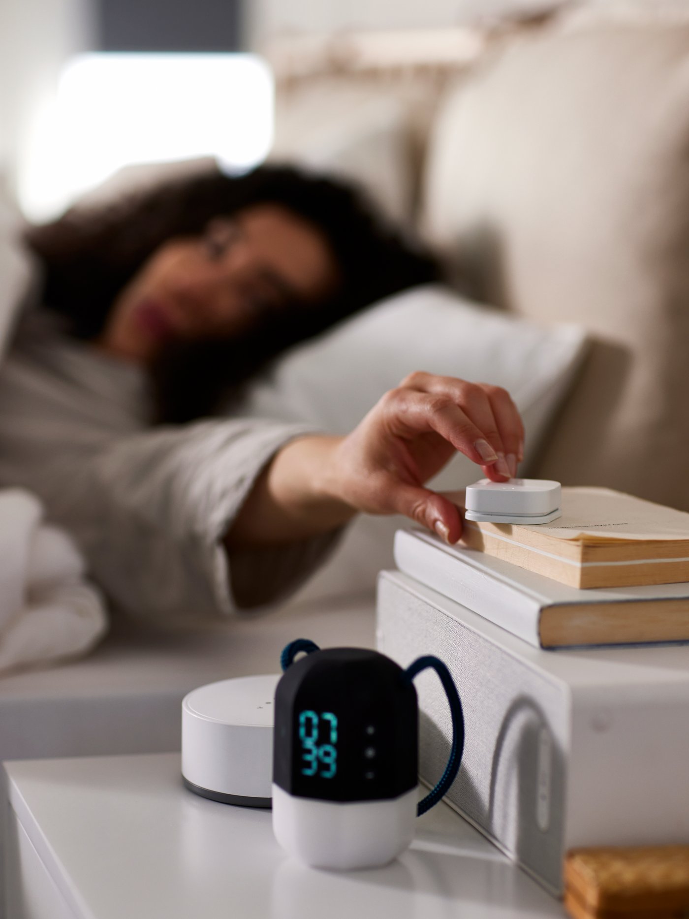 A woman lying in bed pushing a TRÅDFRI wireless dimmer on a pile of books on a WiFi speaker on a white bedside table.