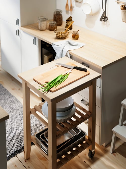 A beige chopping board on a wooden trolley, next to a kitchen cabinet with two jars on the table top.