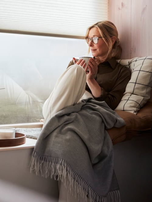Un salon dans lequel on retrouve une personne avec un jeté, une tasse près de coussins, un vase avec des fleurs, un bol, un plateau, une lampe et des stores.