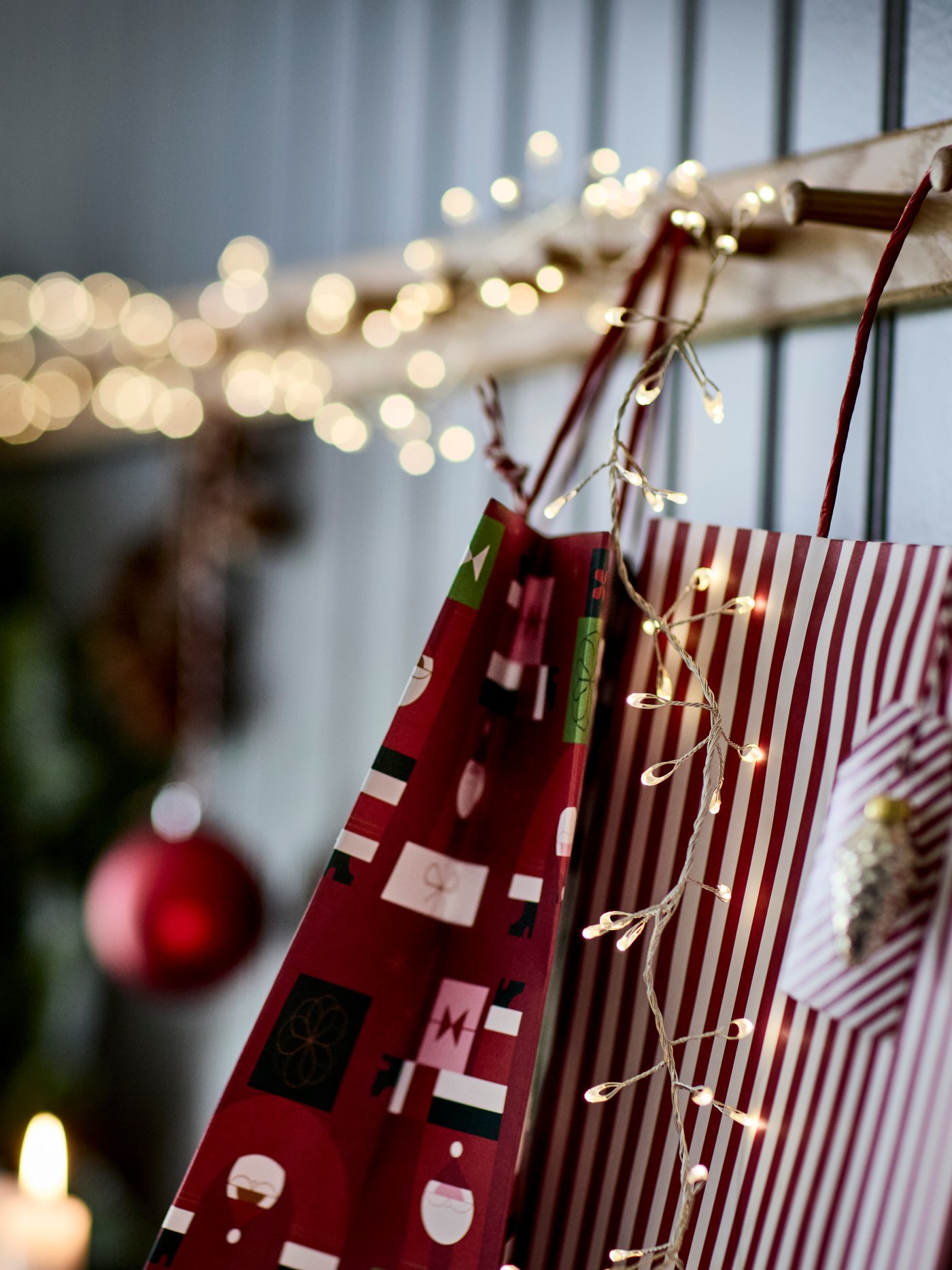 Två juliga presentpåsar hänger på en trähylla med ljusslinga. En har rött mönster, den andra rödvita ränder. Juldekorationer och grönska i bakgrunden skapar en mysig stämning.
