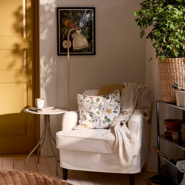 Corner of a room with a white ROCKSJÖN armchair with a side table and reading lamp, and a green plant on the other side.
