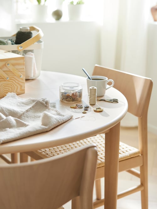 On an extendable table, surrounded by chairs, there is a RISATORP basket, a white IKEA 365+ mug and sewing accessories.