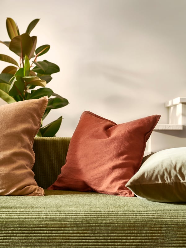 Eine Grünpflanze steht hinter einem JÄTTEBO Sofa mit Kissen in einem goldbraunen und einem olivgrünen SANELA Kissenbezug.