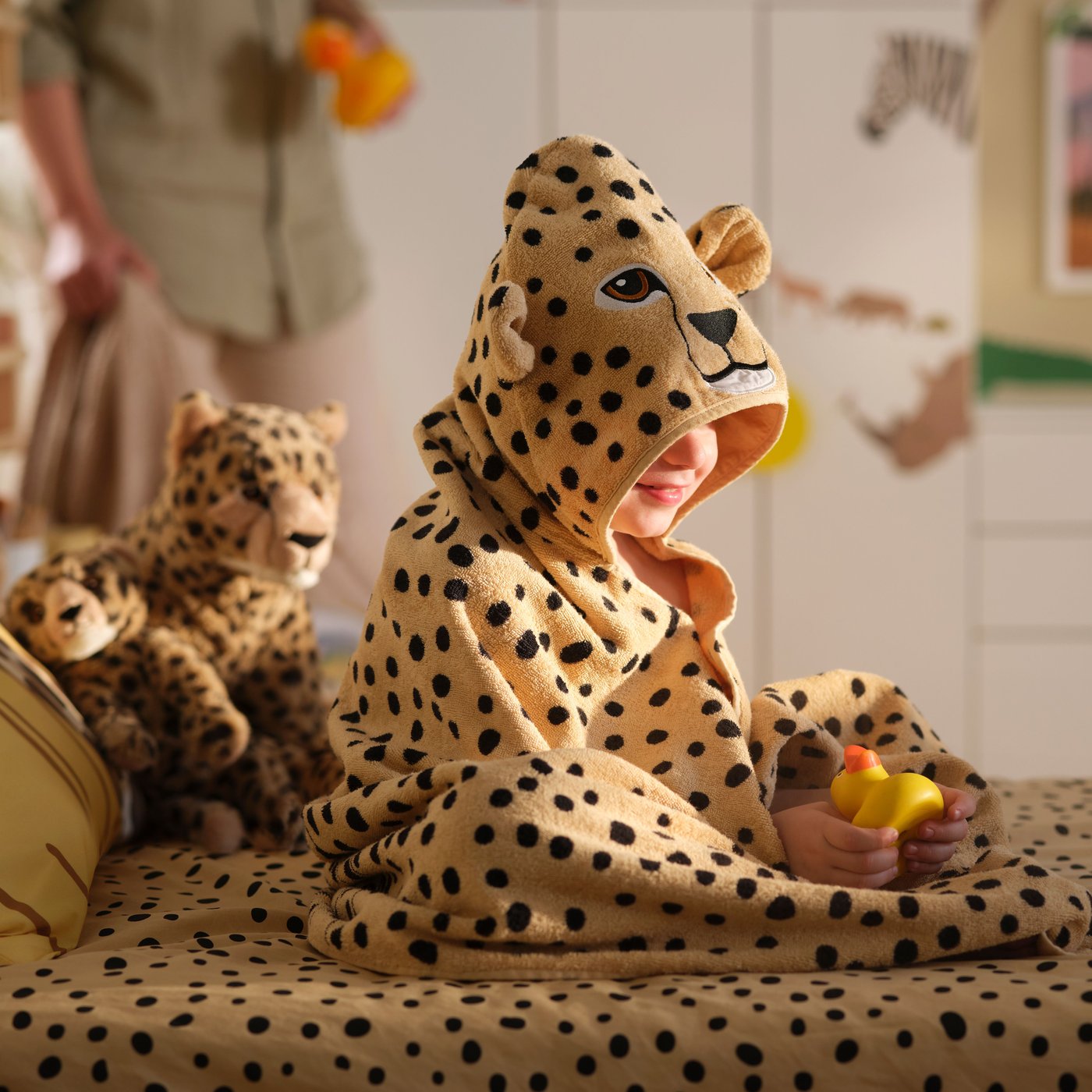 A child in a SANDLÖPARE towel with hood with cheetah pattern sits on a bed surrounded by soft toy cheetahs and a rubber duck.