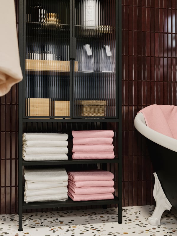 A MOSSJÖN glass-drawer cabinet with 2 doors, filled with white and pink towels and storage boxes, next to a bathtub.