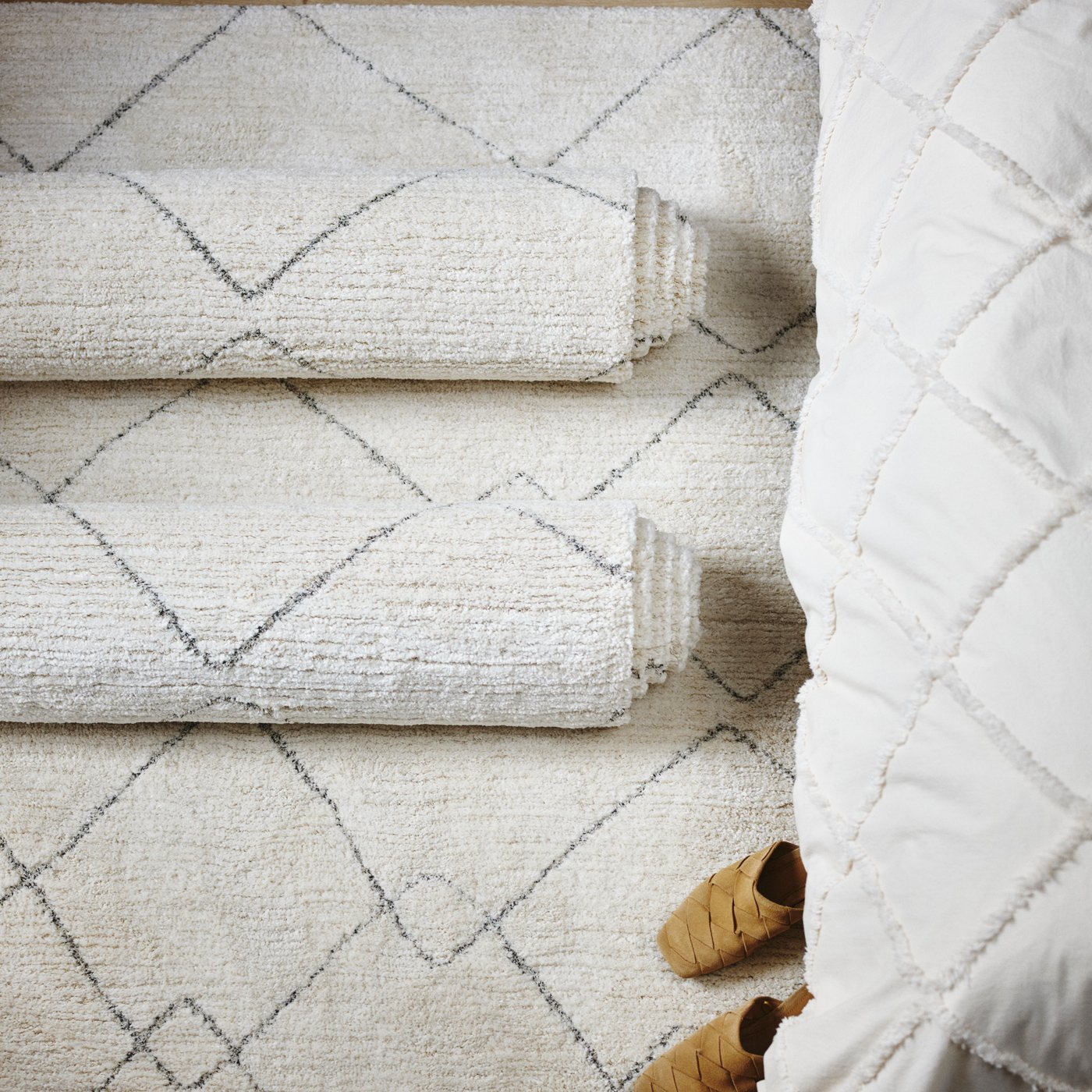 An overhead shot of a plush, off-white rug with a diamond pattern, partially rolled up to show its texture next to a white comforter.