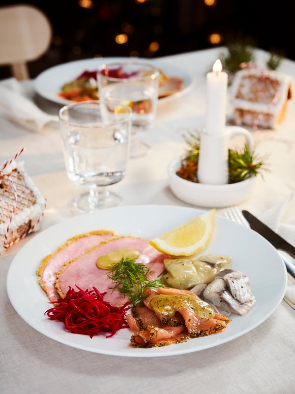 Valkoisella lautasella on erilaisia jouluruokia kuten SJÖRAPPORT-graavilohta, SILL DILL -tillisilliä, SILL SENAP -sinappisilliä sekä kinkkua jonka päällä on SÅS SENAP & DILL -graavilohikastiketta.