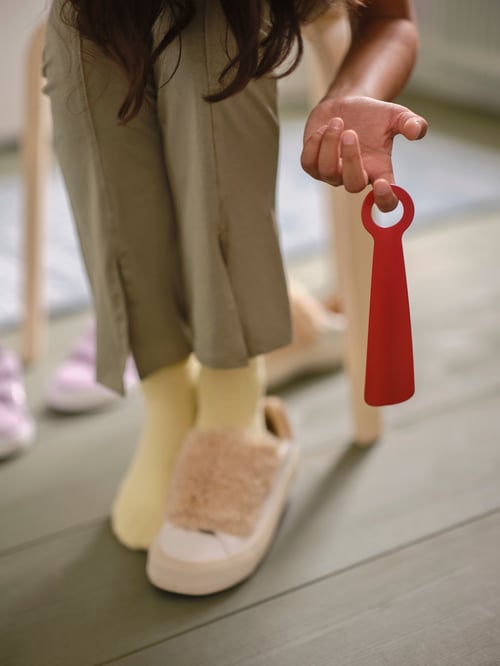 Focused shot of a red SNÖSKYFFEL shoehorn held by one finger by a person wearing yellow socks and one pinks shoe.
