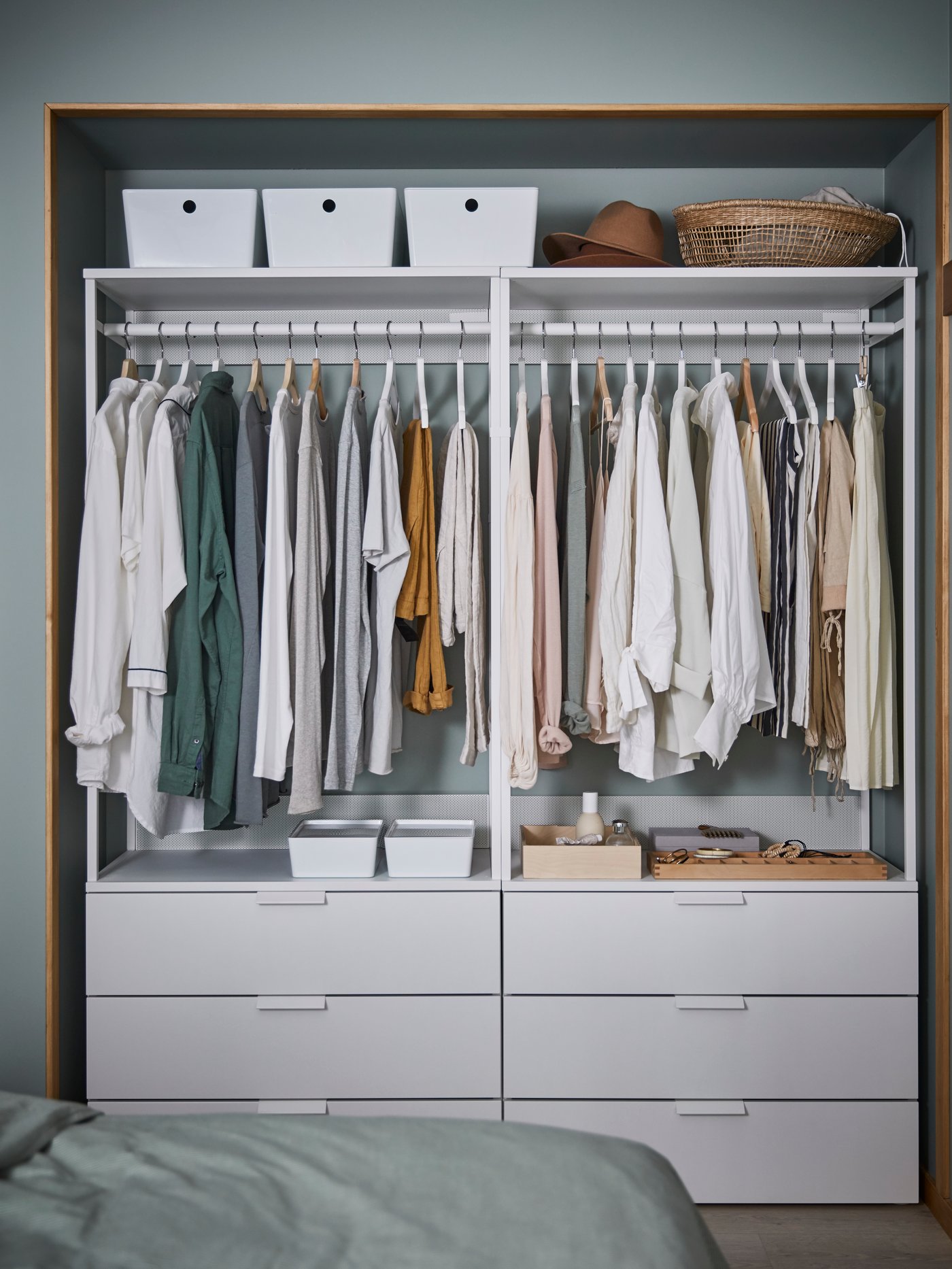 Ambiente de dormitorio con dos armarios blancos con cajones y puertas, con cajas con tapa y varias prendas colgadas.