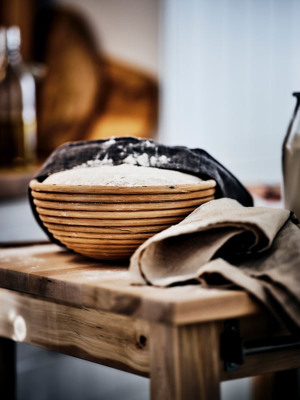 A wooden table with a proofing basket filled with bread dough on it. A tea towel is half pulled across the dough.