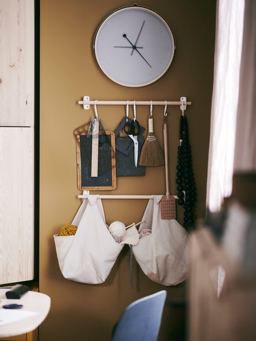 A modern workspace that features a wooden rail with white hangers holding several items, two beige bags, and a wall clock above.