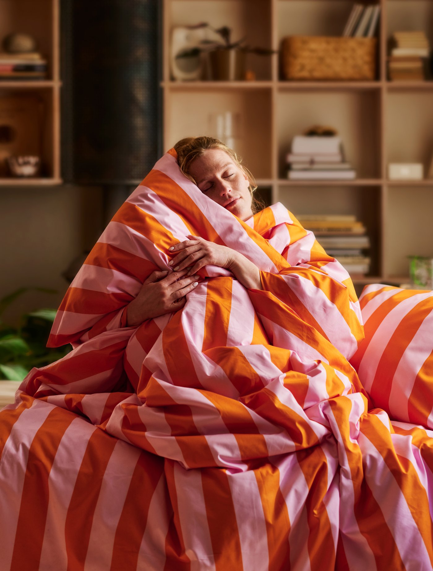 En person med slutna ögon sitter upp i sängen och kramar ett stort, fluffigt täcke med breda ränder i rosa och orange.