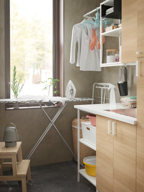 A DÄNKA ironing board next to a drying rack, a laundry, and a step stool with a Bluetooth speaker lamp placed on top. 