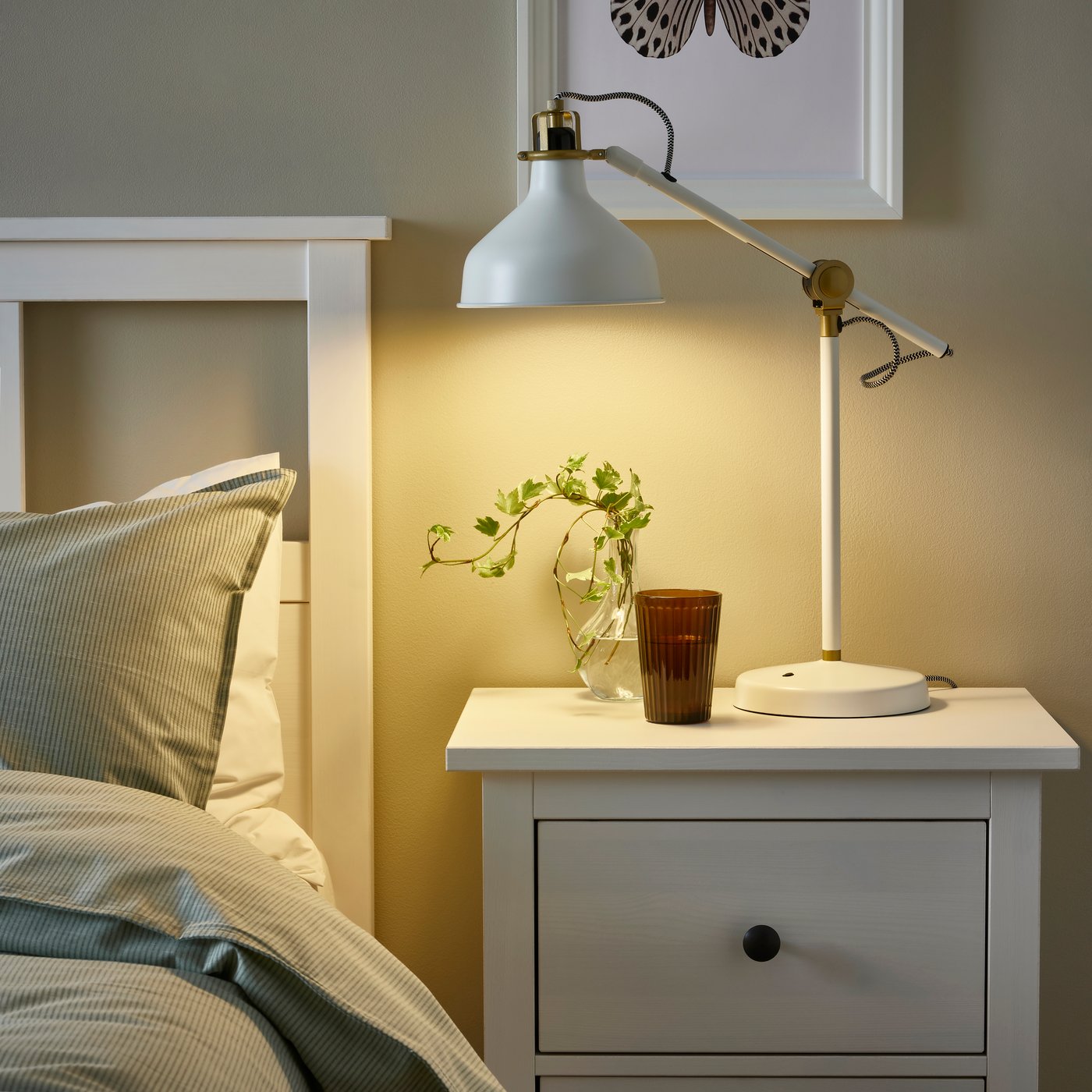 A white RANARP task lamp on a bedside table in a bright bedroom.
