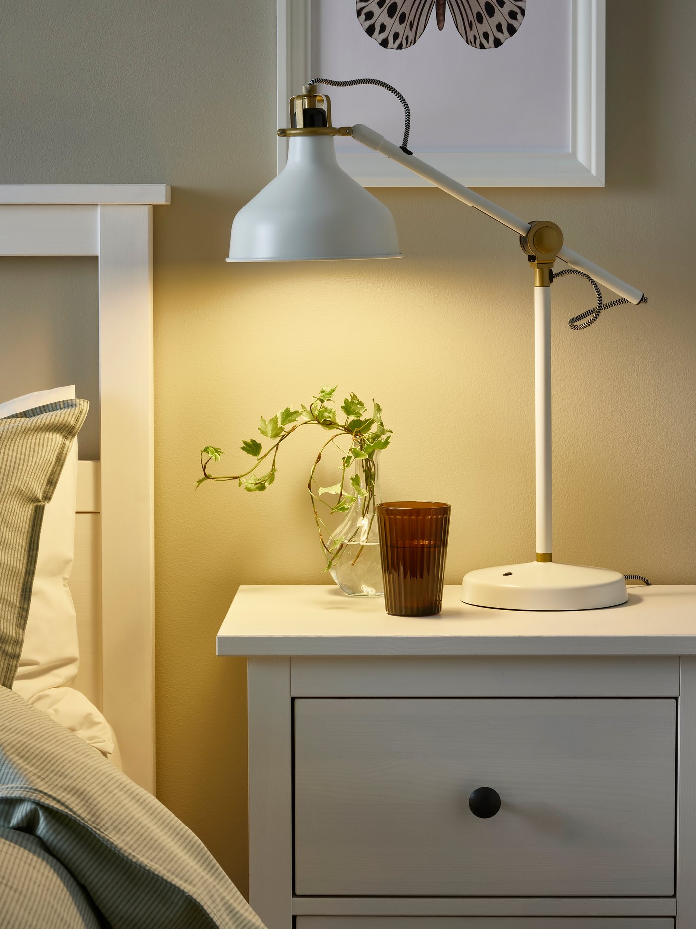 En vit RANARP arbetslampa på ett sängbord i ett ljust sovrum.
