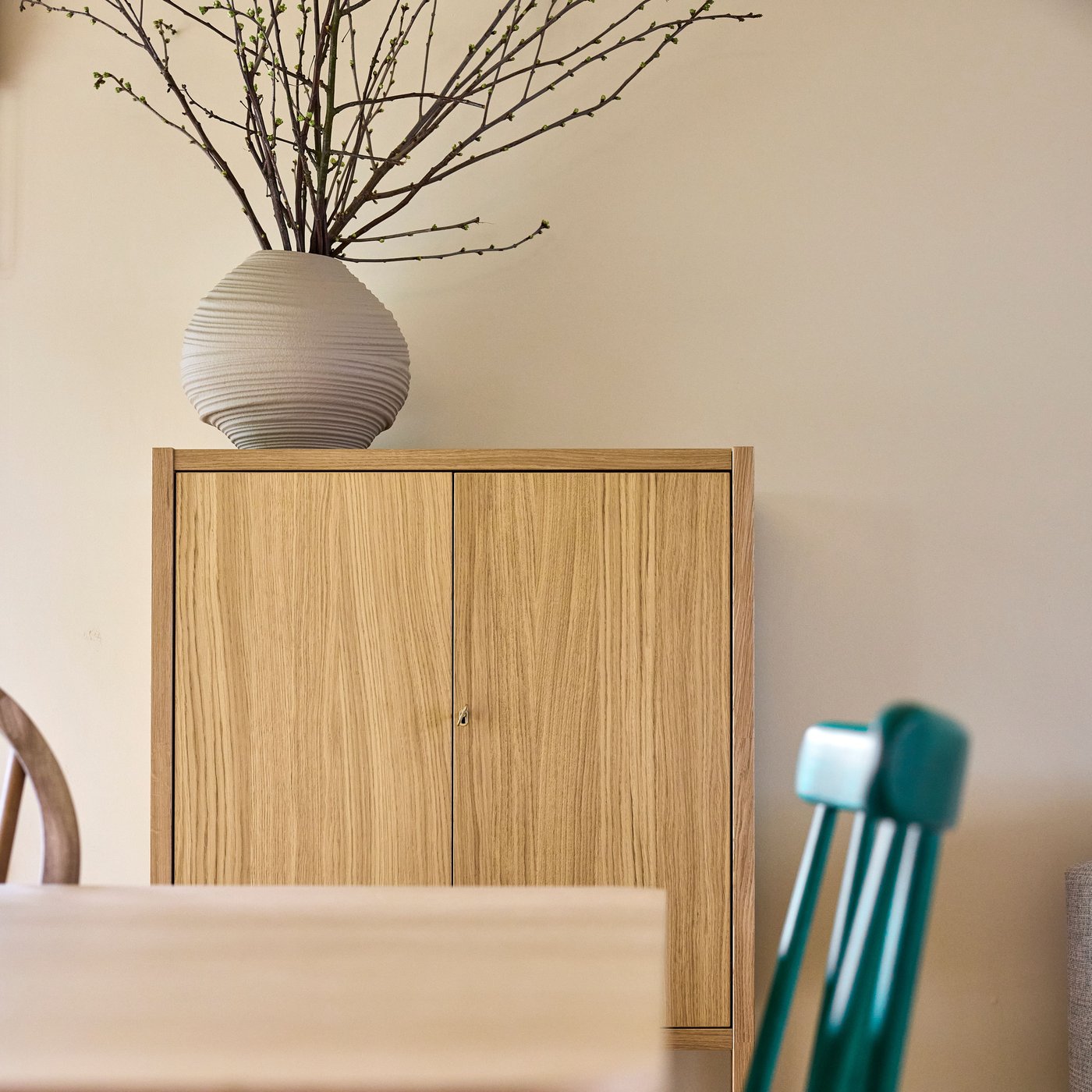Houten kast met twee deuren en decoratieve vaas met takken erboven, in een warme, minimalistische interieursetting.