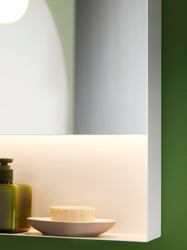 The corner of a white FAXÄLVEN mirror with built-in lighting. An EKOLN soap dish is on the shelf underneath the mirror.