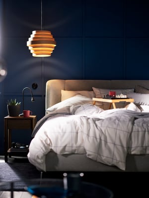 A TUFJORD upholstered bed with two single duvets in BREDVECKLARE bed linen on it and a DYKARKLOCKA pendant lampshade above.