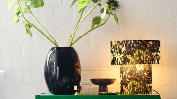 A black vase with green branches placed on a table next to a lamp and a wall featuring gold‑colored artwork.