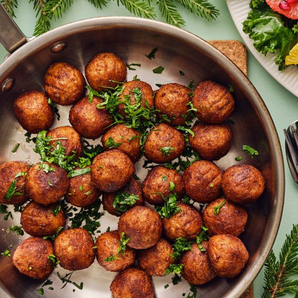 Su una superficie verde chiaro, una padella piena di polpette vegetali HUVUDROLL dorate, guarnite con erbe aromatiche fresche.