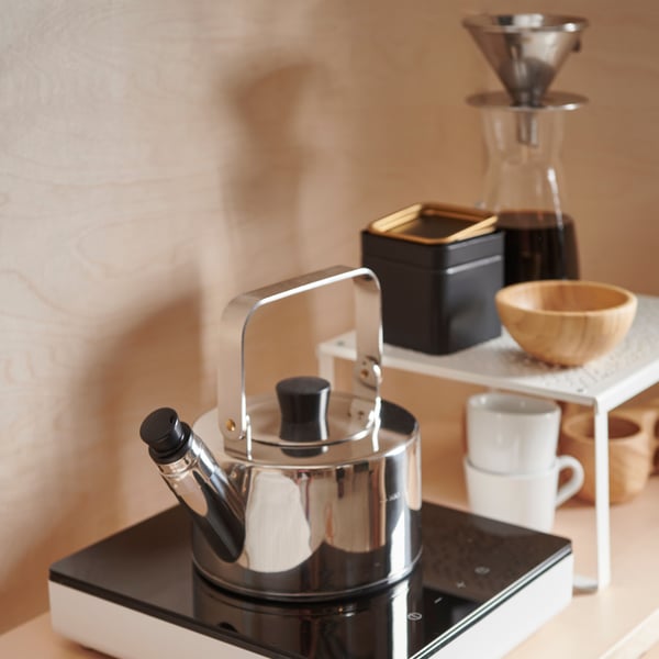 A METALLISK kettle standing on a TILLREDA portable induction hub. In the back, there is a VARIERA shelf insert with IKEA 365+ mugs underneath and a BLANDA MATT bowl on top.