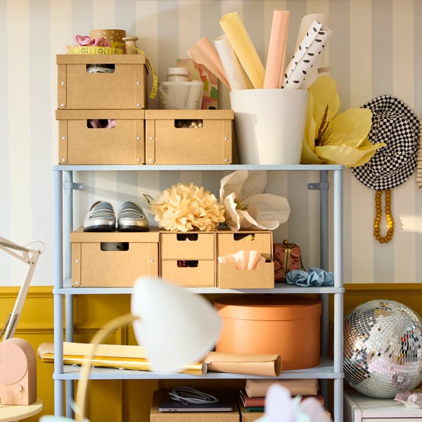 A bright wall with a light grey-blue SÅGMÄSTARE shelving unit, displaying organised boxes and decorative items on shelves.