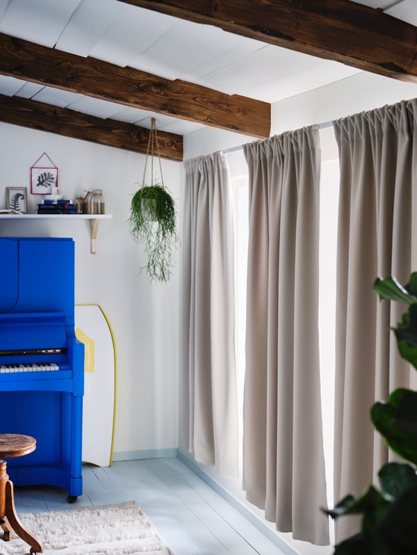 A piano standing beside windows semi-covered by three curtains made from light grey-beige, room darkening MAJGULL fabric.