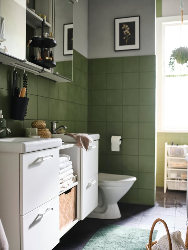 A bathroom with green tiled walls, white ENHET cabinets, two white sinks with drawers and shelves holding white towels.