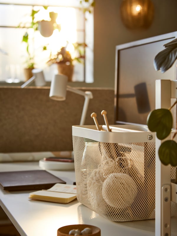 Es ist ein sonniger Arbeitsplatz mit einem RISATORP Korb auf einem Schreibtisch zu sehen, in dem sich Büroutensilien befinden. Auf dem Fensterbrett neben dem Arbeitsplatz stehen verschiedene Pflanzen.