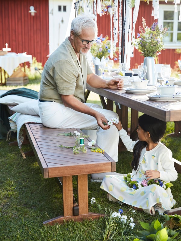En morfar sidder på ved et udendørs bord dekoreret til en havefest, mens et barn sidder nedenunder og er ved at binde en krans af blomster.