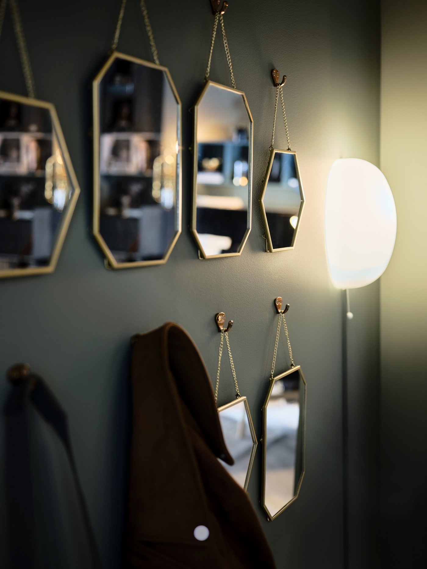 Rows of gold-colour LASSBYN mirrors hang on a green wall amid coats on hooks and a lit wall-mounted lamp.