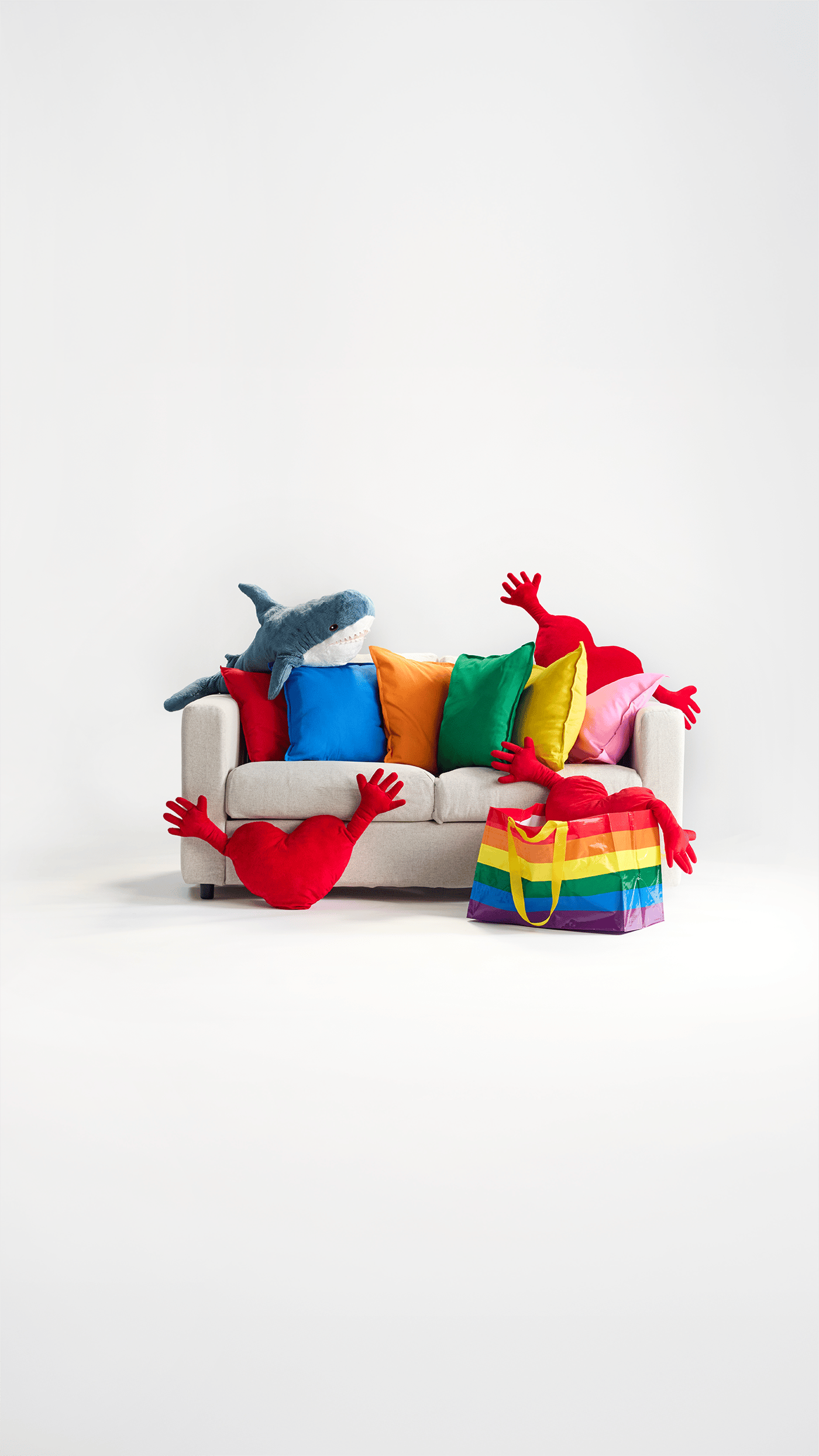 An IKEA sofa covered in rainbow colored products. Rainbow bags, toss pillows, BLAHAJ, and heart-shaped hug pillows.