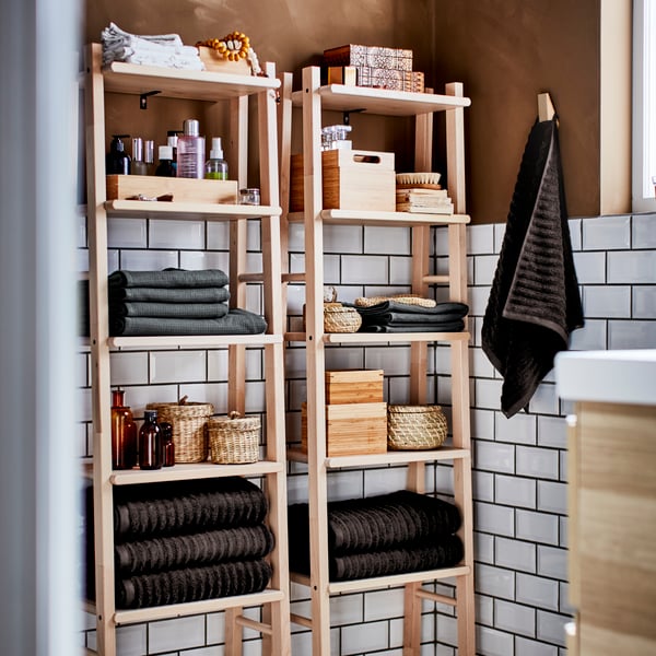 Dos estantes de madera VILTO en un baño con toallas, cajasy accesorios en sus repisas.