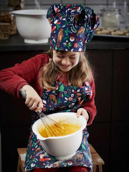 A smiling girl in VINTER 2021 children's hat and apron attacks batter in a white VISPAD mixing bowl with a balloon whisk.