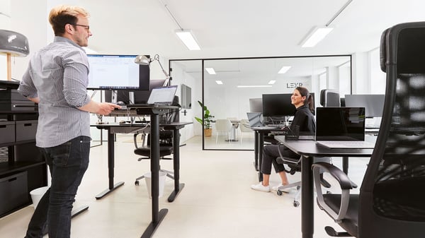 Un espace de bureau avec des bureaux noirs pour s'asseoir ou se tenir debout. Deux collaborateurs discutent.