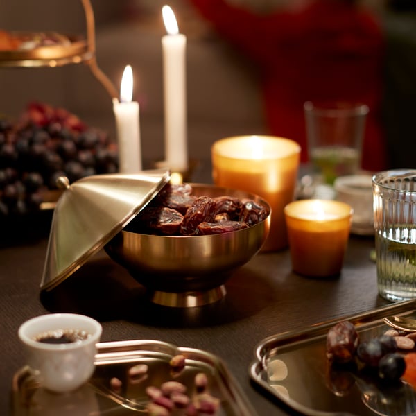 A gold-coloured GOKVÄLLÅ serving bowl filled with dates; next to some GOKVÄLLÅ side plates and a GOKVÄLLÅ tealight holder.