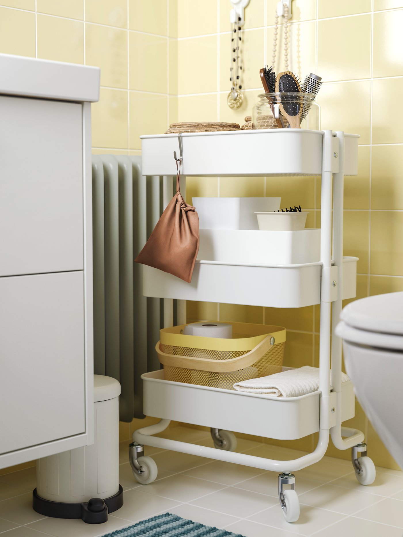 Un carrello RÅSKOG bianco contenente prodotti da bagno è posizionato in un angolo, tra il WC e un mobile per lavabo HAVBÄCK.