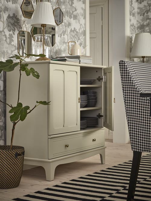 Dining area with a beige cabinet with one open door on which is a table lamp and a watering can, and a chair on a rug.