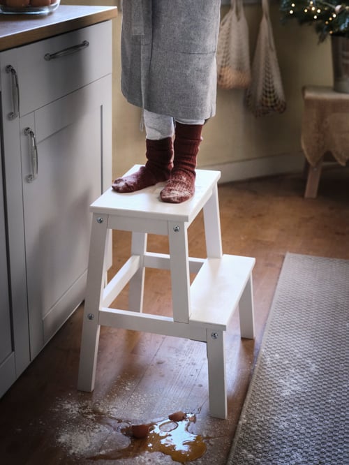 Child wearing apron in kitchen sitting on a sturdy white step stool made of solid wood, next to white kitchen cabinets.