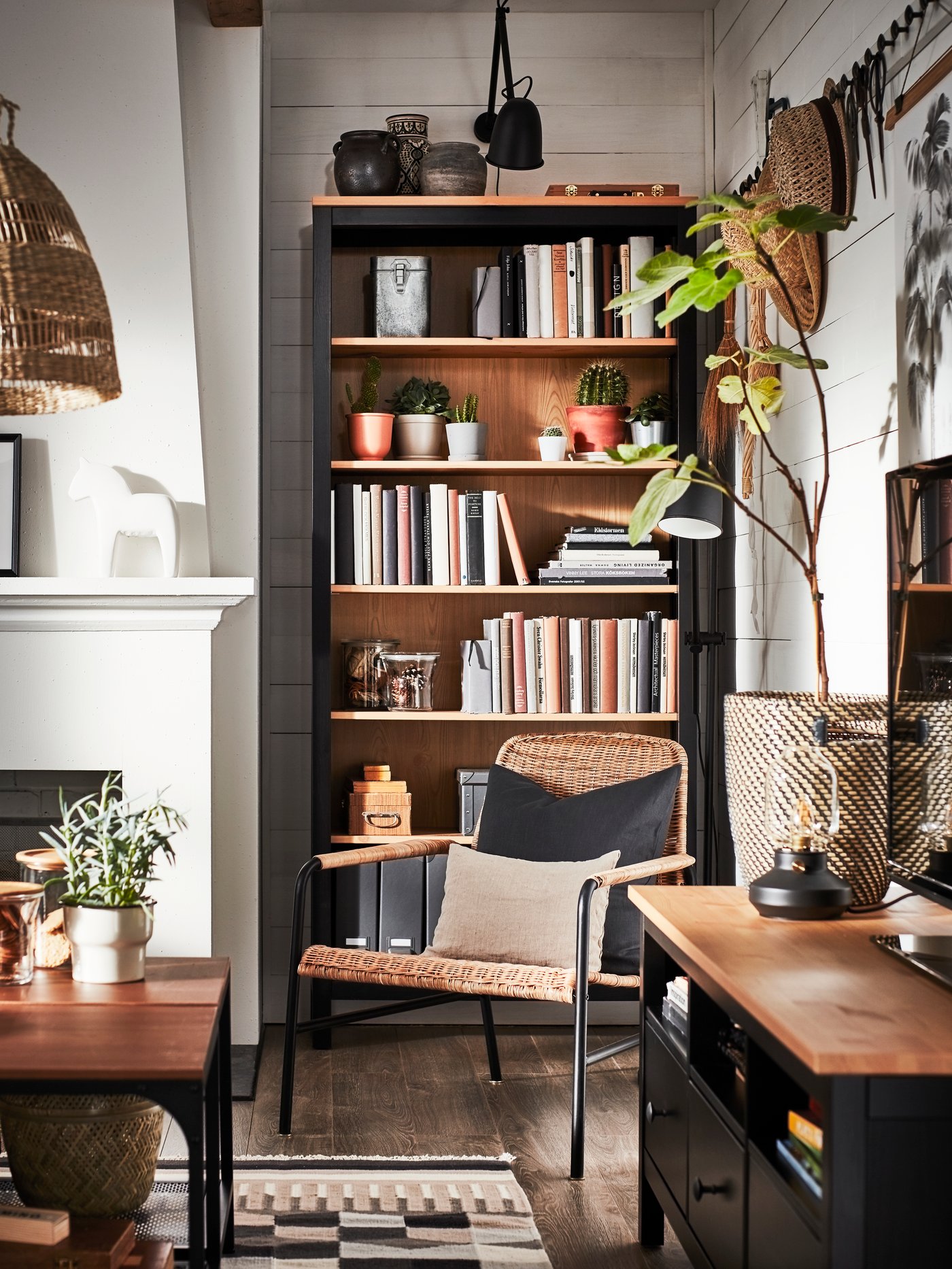 Salón con una librería HEMNES negra con libros y otros objetos. Un sillón de ratán delante con dos cojines.