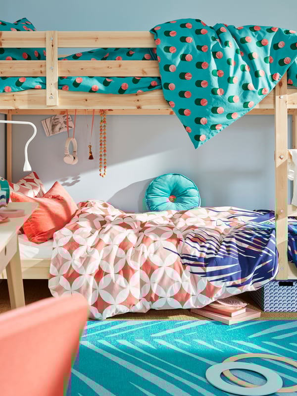 Un camarote MYDAL de madera natural apoyado en una pared celeste claro, las camas tienen ropa de cama de colores GRACIÖS y se ven desordenadas con objetos en uso. 