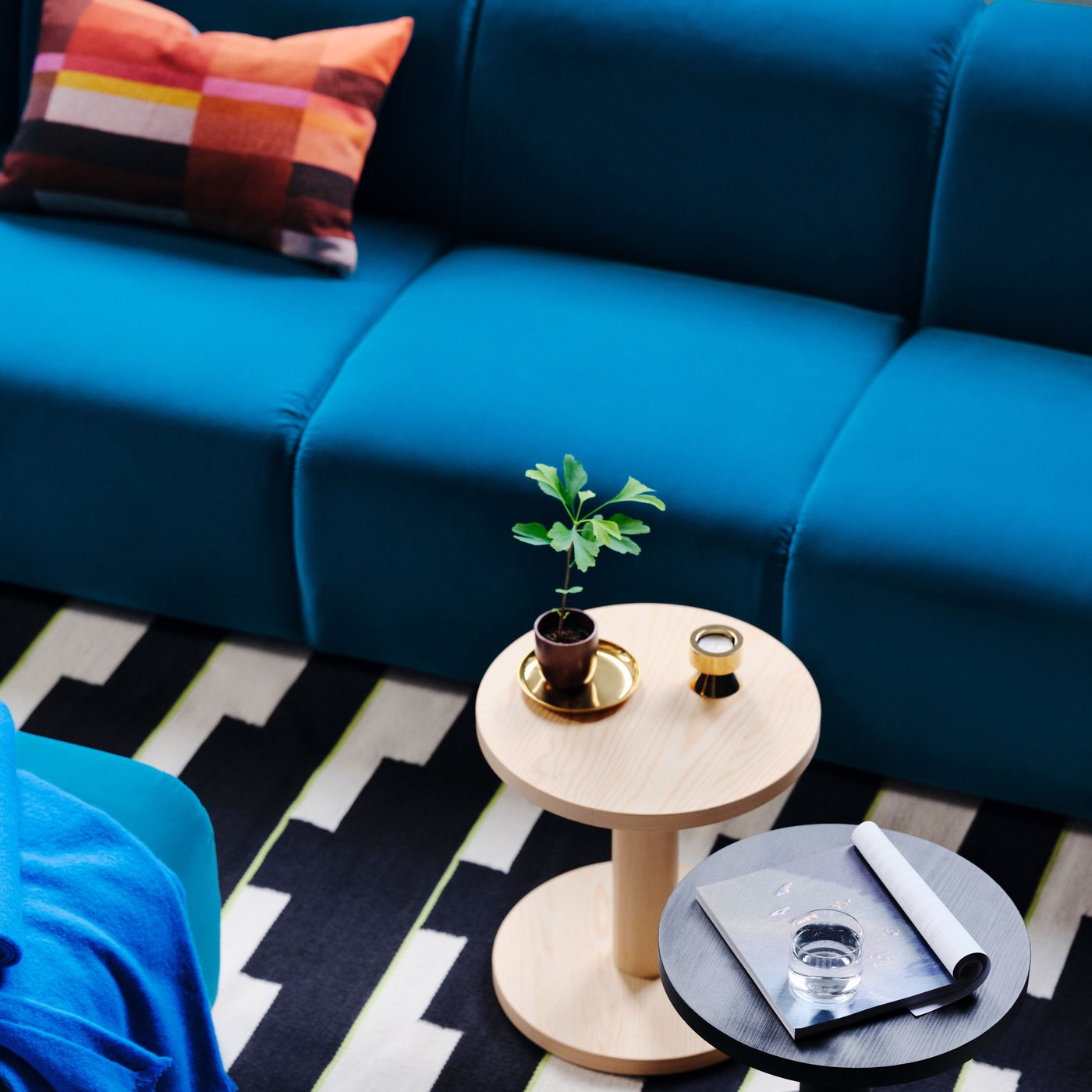 Blå STOCKHOLM soffa med färgglad kudde, två runda bord med växt, ljus, glas vatten och anteckningsbok – en modern och harmonisk inredning.
