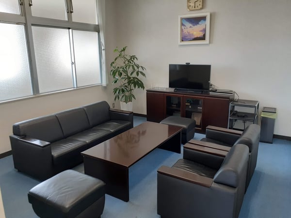 黒いソファセットと木製ローテーブルを置いた応接スペース。テレビ台と観葉植物があり、壁に風景画が掛かっている。