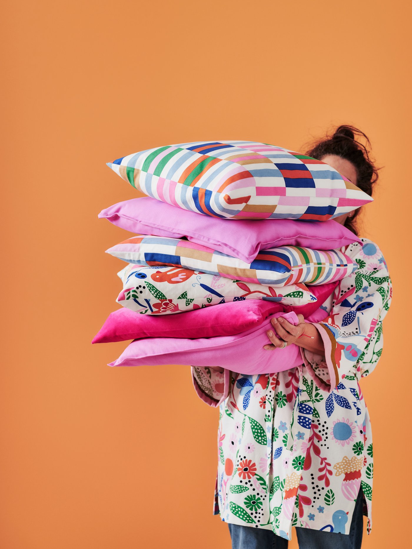 En person håller i flera färgglada, mönstrade kuddar mot en orange bakrgund.