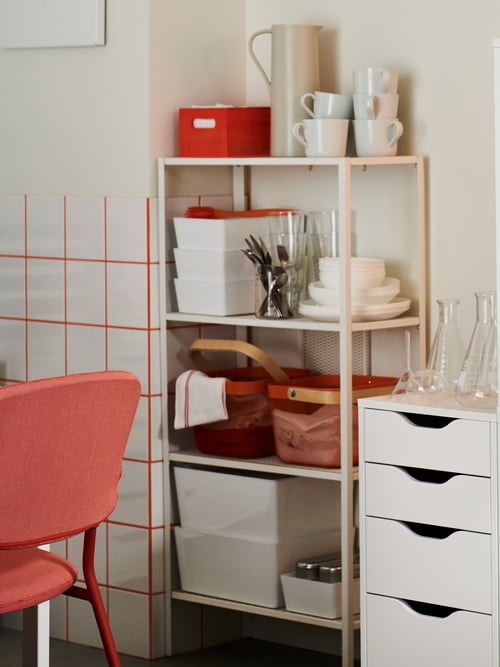 An open shelving unit in white holding various kitchen utensils, baskets and containers, placed next to a white drawer unit.