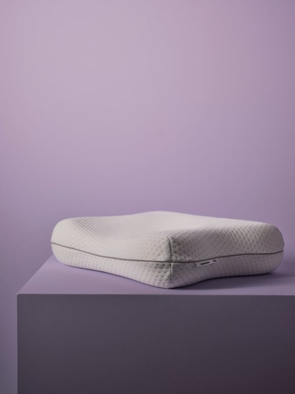 An ISRANUNKEL ergonomic pillow lays flat on a purple pedestal, with a purple wall behind it. No other products are shown.