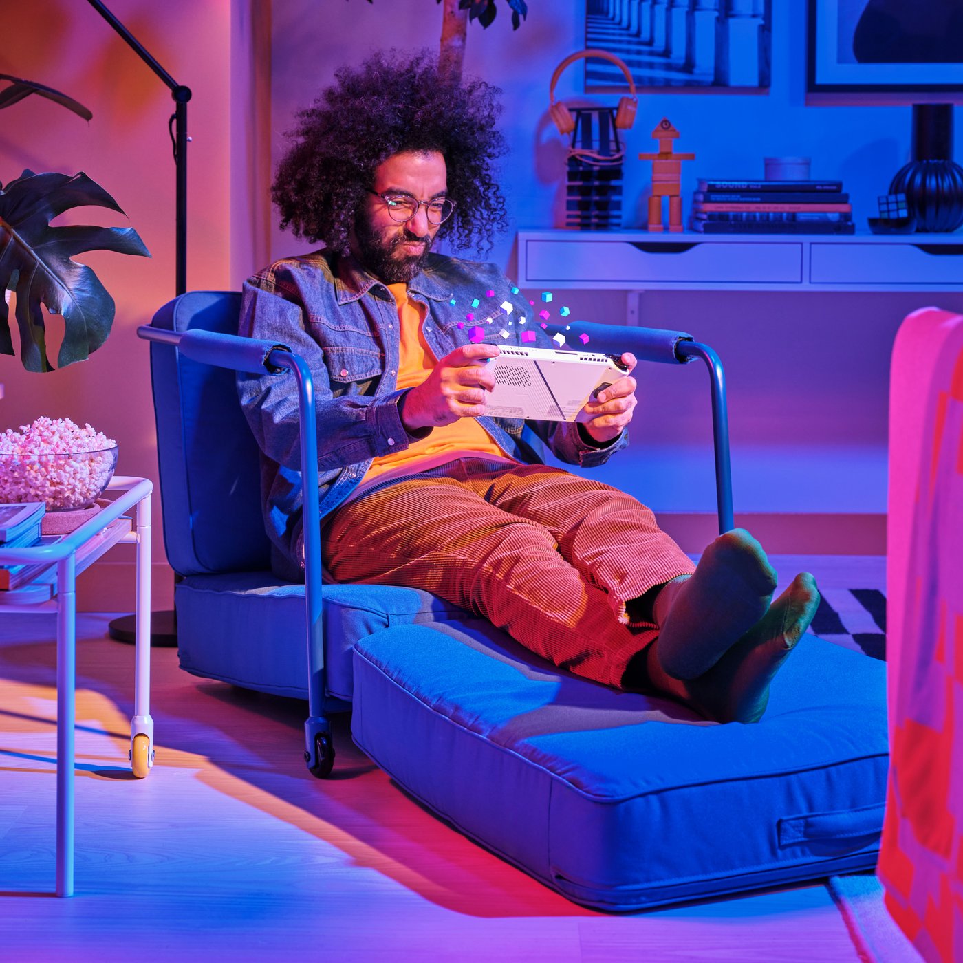 A light ash effect/white BRÄNNBOLL side table on castors and blue BRÄNNBOLL gaming lounge chair. A man is playing games on a console while resting on the gaming lounge chair.