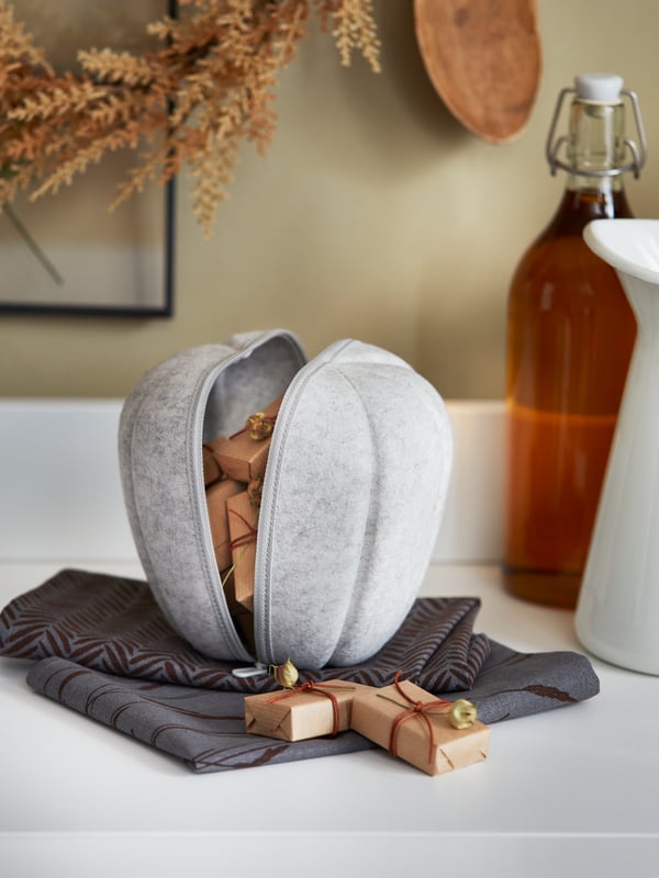 A grey felt HÖSTKVÄLL pumpkin filled with small gifts rests on folded HÖSTKVÄLL cloth napkins on a white HAUGA sideboard.